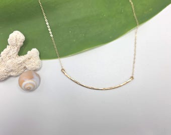 Könnte beinhalten: Eine goldfarbene Halskette mit einer feinen Kette und einem geschwungenen, gehämmerten Metallstab als Anhänger. Der Anhänger hat eine texturierte Oberfläche und ist an beiden Enden mit der Kette verbunden, was eine schlichte Bogenform ergibt.