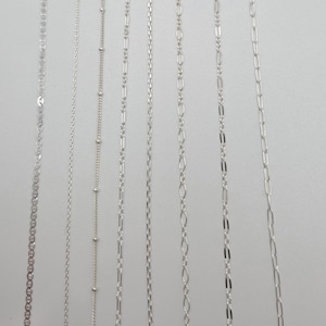 May include: A collection of seven distinct silver-toned metal chains arranged vertically. The assortment includes various link styles such as cable, satellite with small beads, figaro, rope, and elongated oval links, all featuring a polished finish.