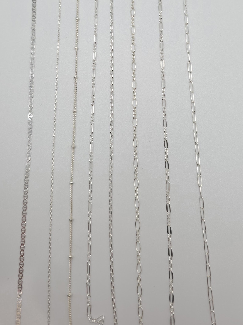 May include: A collection of seven distinct silver-toned metal chains arranged vertically. The assortment includes various link styles such as cable, satellite with small beads, figaro, rope, and elongated oval links, all featuring a polished finish.