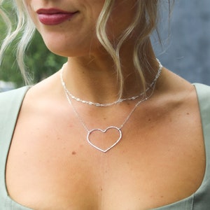 Puede incluir: Un collar de plata con una cadena delicada y un gran colgante de coraz&oacute;n abierto.