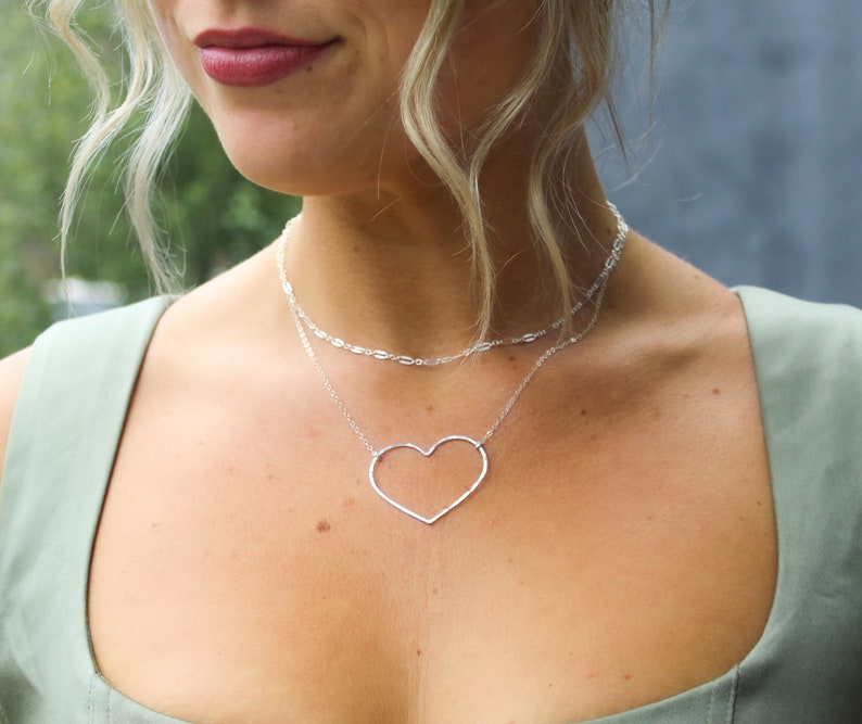 Puede incluir: Un collar de plata con una cadena delicada y un gran colgante de coraz&oacute;n abierto.