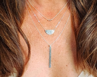 May include: A layered silver necklace with three pendants. The top pendant is a small circle with three beads, the middle pendant is a hammered half-moon shape, and the bottom pendant is a long, hammered bar.