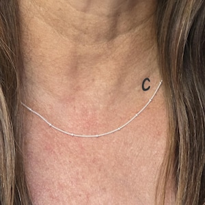 May include: A delicate silver chain necklace featuring evenly spaced small round beads along the length of the chain. The necklace is positioned around the neck, and the letter C is printed in black near the left side of the silver chain.