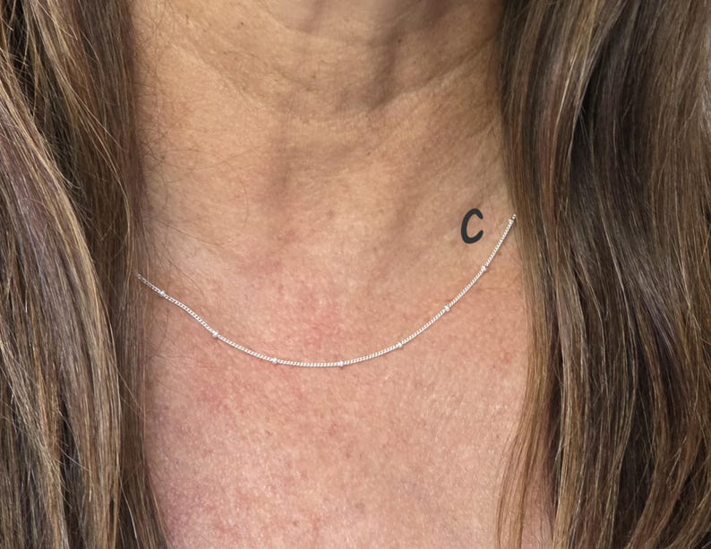 May include: A delicate silver chain necklace featuring evenly spaced small round beads along the length of the chain. The necklace is positioned around the neck, and the letter C is printed in black near the left side of the silver chain.