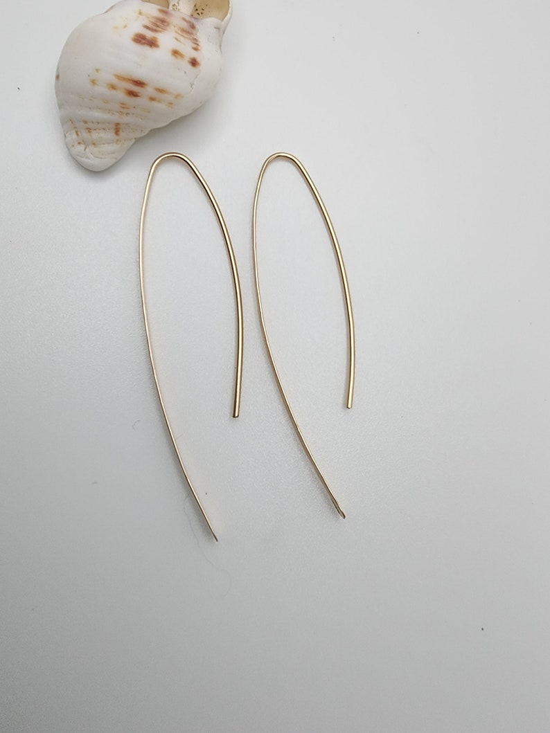 Pode incluir: Um par de brincos de metal em tom dourado com um design de fio longo, fino e em forma de U. Os brincos t&ecirc;m um acabamento liso e polido e uma silhueta curva e alongada que termina em ponta nas extremidades inferiores da estrutura.