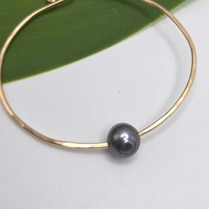 Pode incluir: Uma pulseira circular fina de tom dourado com uma única pérola esférica, lisa e cinzento-escura, centrada ao longo do fio. A banda de metal tem um acabamento polido e um design simples e aberto que forma um círculo completo.