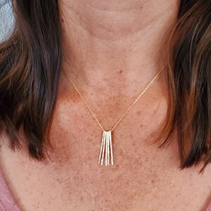K&ouml;nnte beinhalten: Eine goldene Halskette mit einem Anh&auml;nger aus mehreren d&uuml;nnen, vertikalen St&auml;ben. Die Halskette wird von einer Person mit braunen Haaren getragen.