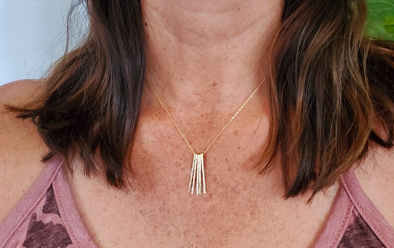 K&ouml;nnte beinhalten: Eine goldene Halskette mit einem Anh&auml;nger aus mehreren d&uuml;nnen, vertikalen St&auml;ben. Die Halskette wird von einer Person mit braunen Haaren getragen.