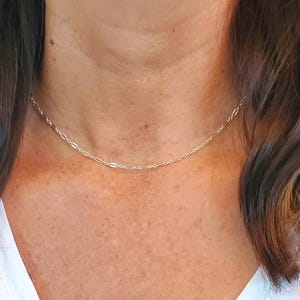 May include: A delicate silver-toned chain necklace featuring a repeating pattern of small, elongated oval links interspersed with tiny circular links. The necklace rests closely against the neck, showcasing a polished, reflective metallic finish.