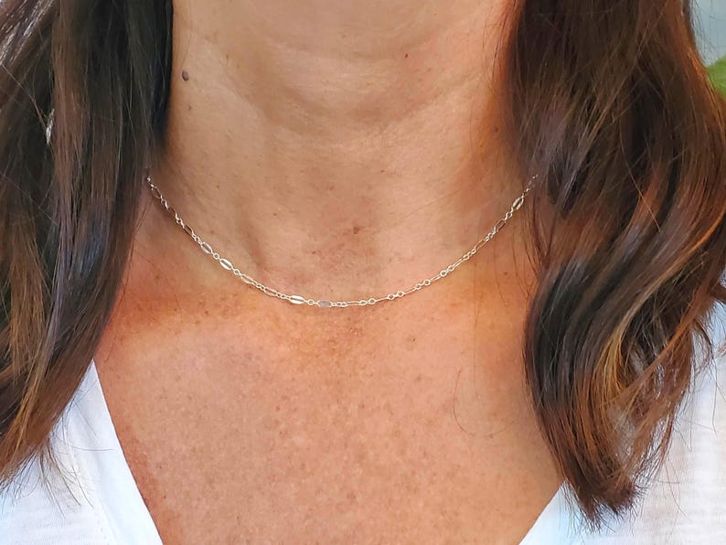 May include: A delicate silver-toned chain necklace featuring a repeating pattern of small, elongated oval links interspersed with tiny circular links. The necklace rests closely against the neck, showcasing a polished, reflective metallic finish.