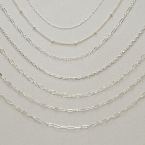 May include: A collection of seven distinct silver-toned metal chains arranged in a cascading arc. The assortment includes various link styles such as delicate cable chains, satellite chains with small beads, and elongated oval link chains.