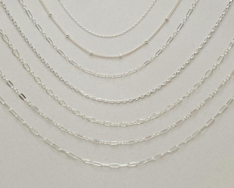 May include: A collection of seven distinct silver-toned metal chains arranged in a cascading arc. The assortment includes various link styles such as delicate cable chains, satellite chains with small beads, and elongated oval link chains.