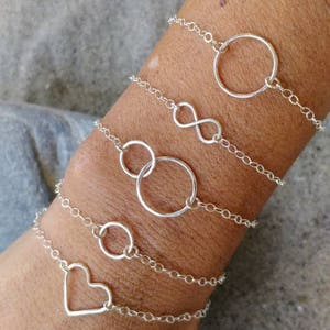 Puede incluir: Un conjunto de cuatro pulseras de plata con diferentes dijes. Los dijes incluyen un círculo, un símbolo de infinito, dos círculos entrelazados y un corazón.
