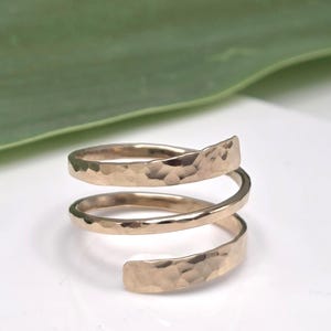 May include: A gold-toned metal ring featuring a spiral design with two full wraps. The surface of the metal band has a distinct hammered texture, creating a series of small, reflective indentations across the entire length of the coiled band.