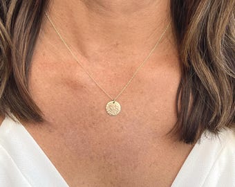 May include: A gold-toned necklace featuring a delicate chain and a small, circular pendant with a textured, hammered surface. The pendant hangs centrally from the chain, which rests against the collarbone area, showcasing a metallic finish.