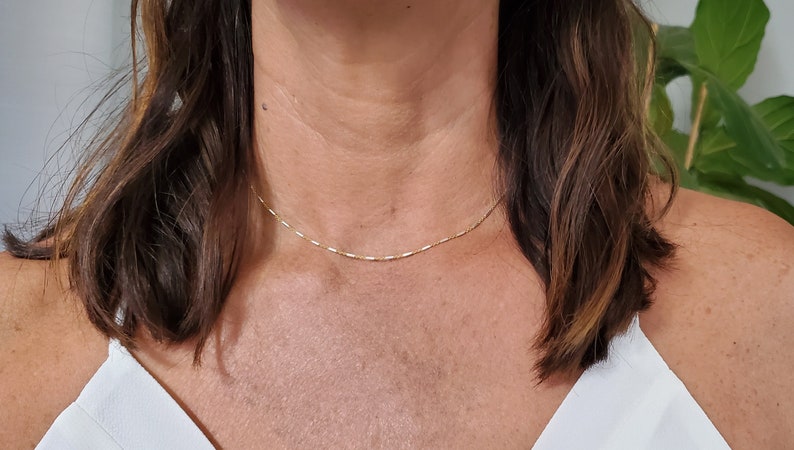 May include: A delicate gold chain necklace worn by a person with brown hair. The chain is thin and has a simple design.