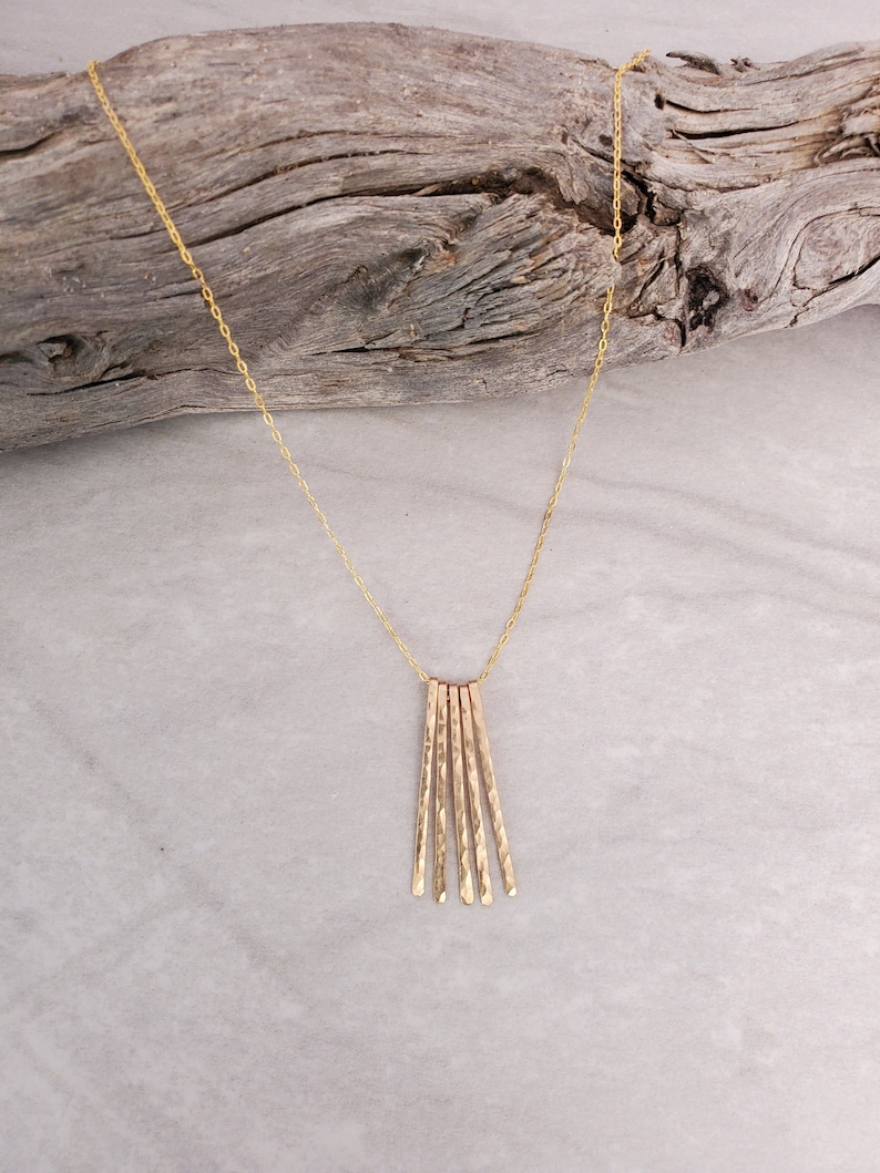 K&ouml;nnte beinhalten: Eine goldene Halskette mit einem Anh&auml;nger aus mehreren d&uuml;nnen, geh&auml;mmerten Goldst&auml;ben. Die St&auml;be sind f&auml;cherf&ouml;rmig angeordnet und h&auml;ngen an einer zarten goldenen Kette.