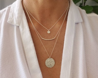 May include: A layered silver necklace with three pendants: a small circle, a thin bar, and a large hammered circle.