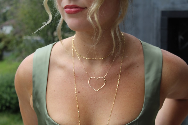 Puede incluir: Una mujer con un top verde sin mangas y un collar de oro de varias capas con un colgante en forma de coraz&oacute;n.