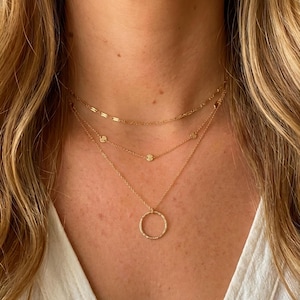 May include: A three-layered gold necklace set featuring a top chain with elongated rectangular links, a middle chain with small circular accents, and a bottom chain with a textured circular pendant. The jewelry is crafted from polished gold metal.