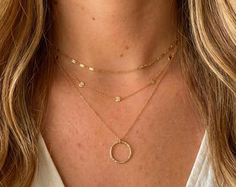 May include: A three-layered gold necklace set featuring a top chain with elongated rectangular links, a middle chain with small circular accents, and a bottom chain with a textured circular pendant. The jewelry is crafted from polished gold metal.