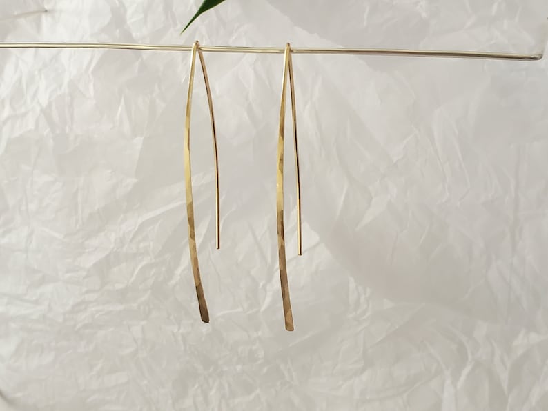 Pode incluir: Um par de brincos dourados longos e finos com um design em forma de V e um acabamento martelado e texturizado. Os brincos est&atilde;o suspensos num fio horizontal fino, exibindo a sua forma met&aacute;lica alongada, curva e superf&iacute;cie dourada.