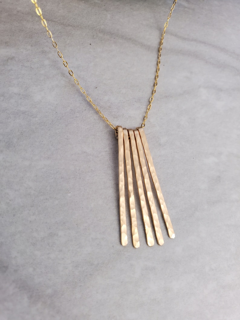 K&ouml;nnte beinhalten: Eine goldene Halskette mit einem geh&auml;mmerten Metallanh&auml;nger mit f&uuml;nf d&uuml;nnen, vertikalen St&auml;ben.