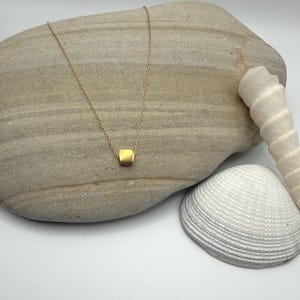 May include: A delicate gold-tone necklace featuring a single, small, smooth square-shaped pendant centered on a thin chain. The necklace is displayed against a large, smooth, light gray stone and a white, ridged, spiral-shaped seashell.
