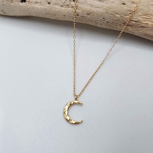 May include: A gold-toned necklace featuring a delicate chain and a small, crescent moon pendant. The moon charm has a textured, hammered surface that reflects light, suspended from a thin, simple link chain that hangs in a gentle curve.