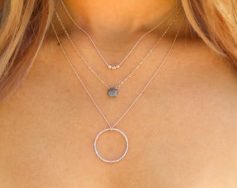 May include: A layered silver necklace with a circle pendant, a small stone pendant, and a row of small beads.