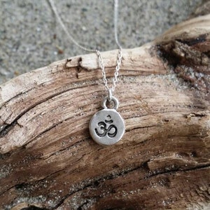 May include: A silver necklace with a small round pendant featuring the Om symbol. The necklace is displayed on a piece of driftwood.