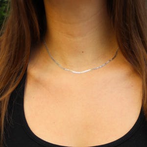 May include: A thin, silver-colored herringbone chain necklace resting against the neck. The necklace features a flat, woven metal link design that creates a smooth, reflective surface. The chain sits close to the base of the throat in a simple style.