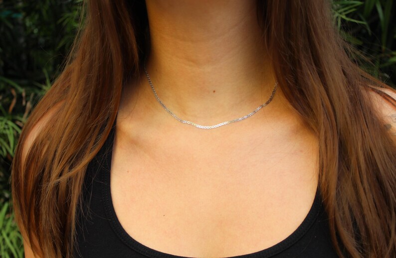 May include: A thin, silver-colored herringbone chain necklace resting against the neck. The necklace features a flat, woven metal link design that creates a smooth, reflective surface. The chain sits close to the base of the throat in a simple style.