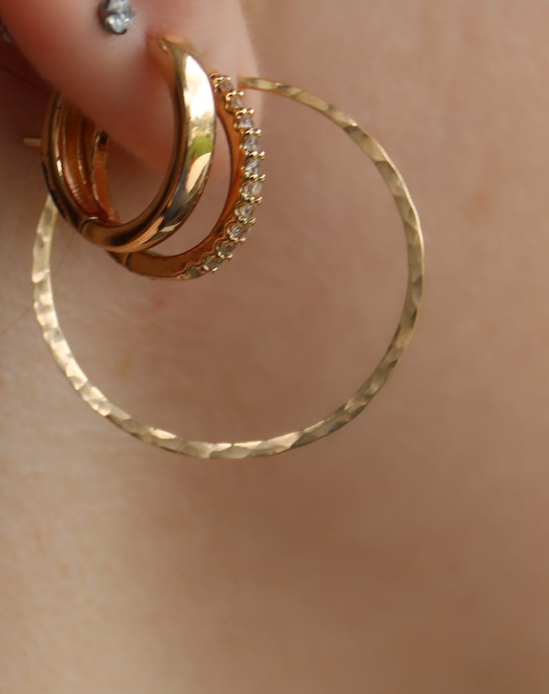 May include: Gold hoop earrings with a hammered texture. The larger hoop is plain, while the smaller hoop has a row of clear cubic zirconia stones.