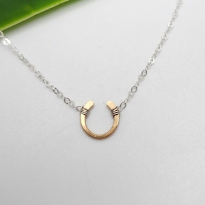 May include: A silver chain necklace with a gold horseshoe pendant. The horseshoe is made of thick wire and has a simple, minimalist design.