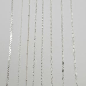 May include: Seven distinct silver-toned metal chains arranged vertically. The collection includes a mariner chain, a thin cable chain, a satellite chain with small beads, a figaro chain, a rope chain, an oval link chain, and a long rectangular link chain.