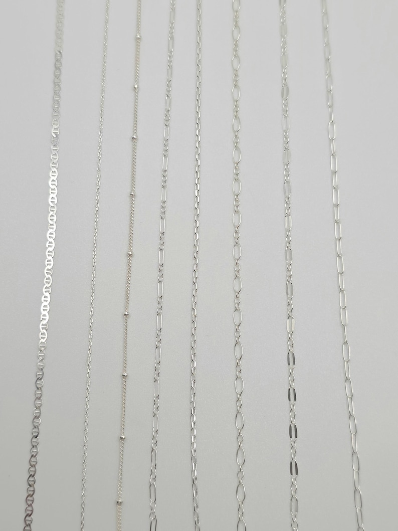 May include: Seven distinct silver-toned metal chains arranged vertically. The collection includes a mariner chain, a thin cable chain, a satellite chain with small beads, a figaro chain, a rope chain, an oval link chain, and a long rectangular link chain.