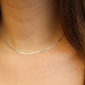 May include: A thin, silver-toned chain necklace featuring a delicate mariner link design. The necklace rests closely against the neck, showcasing a polished metallic finish with uniform, oval-shaped interlocking links that create a subtle, refined appearance.