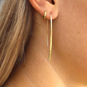 Pode incluir: Um brinco de argola dourado e um brinco longo e fino de fio dourado martelado. O brinco de fio apresenta um design de barra plana e alongada com um acabamento martelado texturizado que capta a luz, pendendo verticalmente do l&oacute;bulo da orelha.