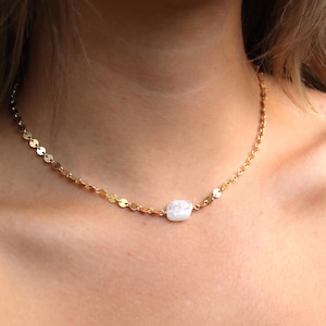 Puede incluir: Un collar de cadena en tono dorado que presenta una serie de pequeños discos metálicos planos y circulares unidos entre sí. El collar está centrado con una sola cuenta de piedra blanca, opaca e irregular, situada en la parte delantera.