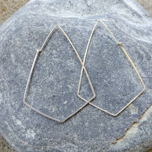 May include: A pair of silver geometric hoop earrings with a diamond shape. The earrings are on a gray rock.
