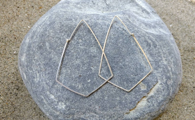 May include: A pair of silver geometric hoop earrings with a diamond shape. The earrings are on a gray rock.