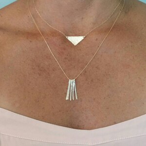 K&ouml;nnte beinhalten: Eine goldene Halskette mit einem Dreiecksanh&auml;nger und einer baumelnden Kette aus kleinen, geh&auml;mmerten St&auml;ben.
