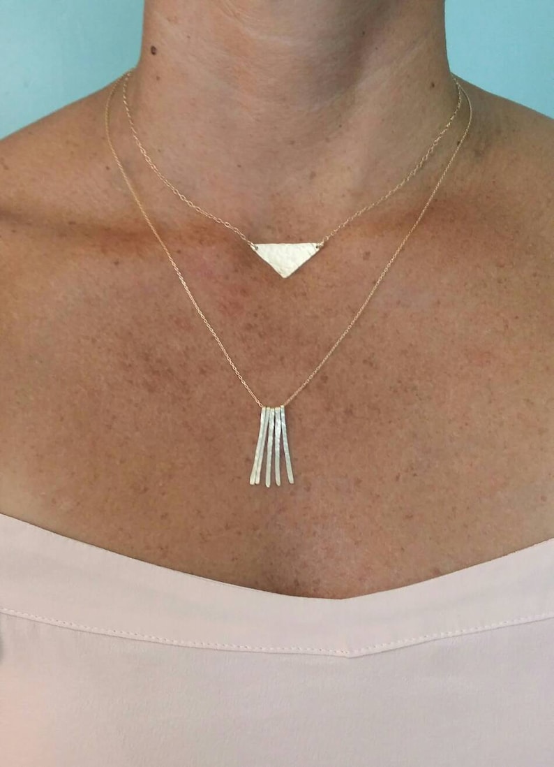 K&ouml;nnte beinhalten: Eine goldene Halskette mit einem Dreiecksanh&auml;nger und einer baumelnden Kette aus kleinen, geh&auml;mmerten St&auml;ben.