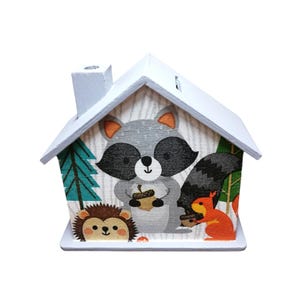 Könnte beinhalten: Ein Sparschwein aus Holz in Form eines Hauses mit hellblauer Lackierung. Die Vorderseite des Hauses zeigt eine bunte Illustration eines Waschbären, eines Eichhörnchens und eines Igels in einer Waldlandschaft.
