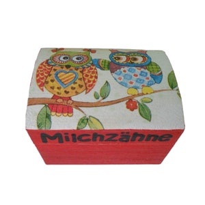 May include: A red wooden box with a white lid featuring two colorful owls perched on a branch. The lid has a floral design and the words "Milchzähne" are printed on the front of the box.
