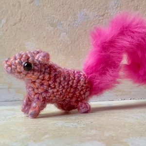 May include: A handmade crochet squirrel with a vibrant pink fluffy tail. The squirrel's body is a mix of pink and orange yarn, with small black eyes. The craft animal is standing on a light-colored surface.