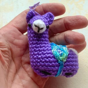 May include: A purple crocheted llama keychain featuring a white muzzle with black stitched details, small black bead eyes, and a shiny blue and green iridescent ribbon saddle. The small stuffed animal is attached to a purple plastic ring.