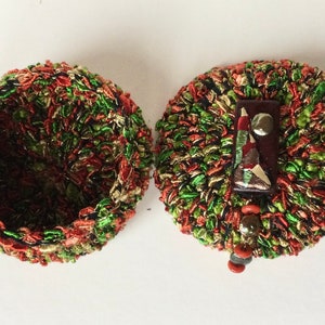 May include: An open, round, handmade container with a matching lid. The container and lid are made of a textured material in shades of green, red, and gold. The lid features a decorative element with a small, colorful design.