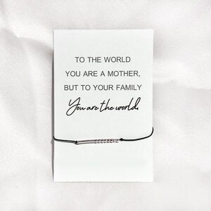 Könnte beinhalten: Eine weiße Karte mit schwarzem Text, der lautet: "Für die Welt bist du eine Mutter, aber für deine Familie bist du die Welt". Ein schwarzes Armband mit silbernen Perlen liegt auf der Karte.
